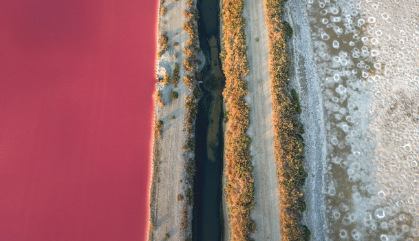 Aerial view of a pink salt evaporation pond separated from a dark water channel and dry mineral covered ground by parallel embankments in Aigues Mortes Occitanie France.