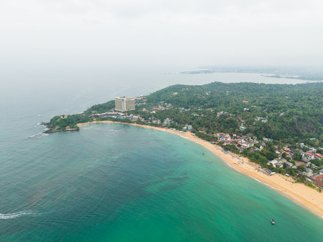 Aerial view of the tropical coastline with a sandy beach turquoise ocean water and lush green hills under an overcast sky in Unawatuna Southern Province Sri Lanka.