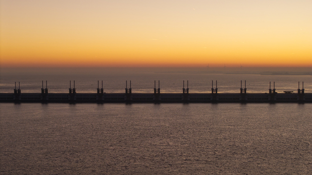 Aerial view of the Oosterscheldekering storm surge barrier with its massive pillars silhouetted against a vibrant orange sunset sky over the calm sea Netherlands.