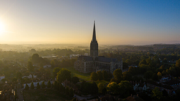 Aerial view of Salisbury Cathedral with its iconic spire rising above the historic city surrounded by lush trees and soft morning mist in Salisbury England United Kingdom. Aerial view of Salisbury Cathedral with its iconic spire rising above the historic city surrounded by lush trees and soft morning mist in Salisbury England United Kingdom.