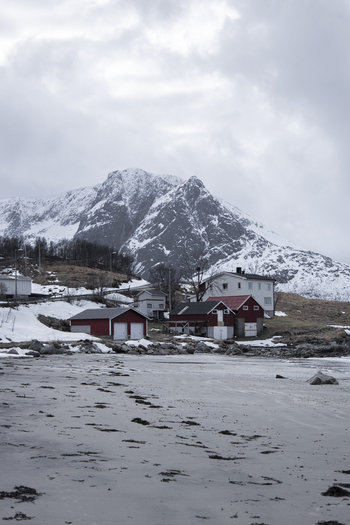 View of traditional red wooden houses on a snowy shoreline with a massive snow covered mountain peak under a cloudy sky in Kvaloya Troms Norway. View of traditional red wooden houses on a snowy shoreline with a massive snow covered mountain peak under a cloudy sky in Kvaloya Troms Norway.
