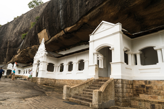 View of the Dambulla Cave Temple featuring white arched entrances built into a massive rock overhang with tourists walking on the stone path Dambulla Central Province Sri Lanka.