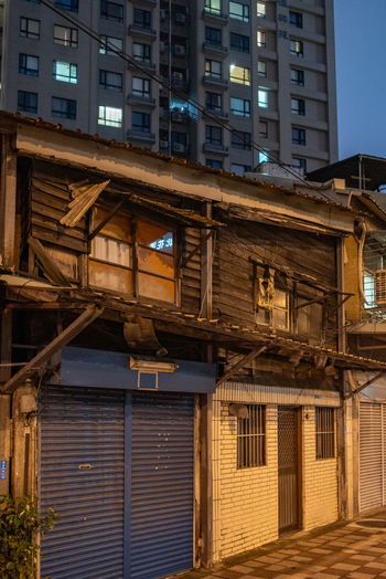 View of an old wooden house with a blue metal shutter standing in contrast to a modern high rise apartment building at dusk in Zhongzheng District Taipei City Taiwan. View of an old wooden house with a blue metal shutter standing in contrast to a modern high rise apartment building at dusk in Zhongzheng District Taipei City Taiwan.