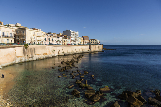 View of sunlight kissing the ancient stone walls and inviting azure waters in a dance of light and shadow Ortigia Sicily Italy.