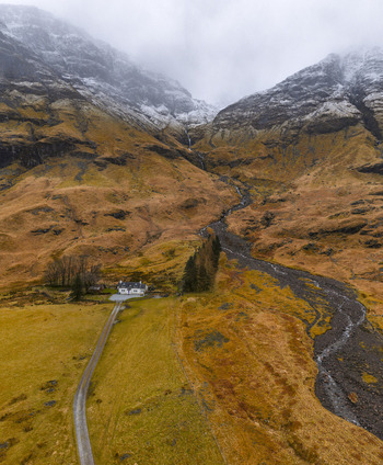 Aerial view of a small white cottage nestled at the base of dramatic snow capped mountains and rugged slopes Glencoe Scotland United Kingdom. Aerial view of a small white cottage nestled at the base of dramatic snow capped mountains and rugged slopes Glencoe Scotland United Kingdom.