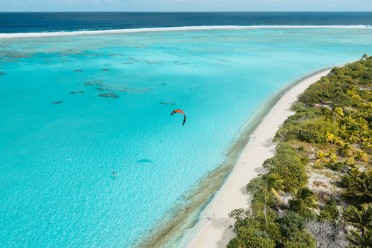 Aerial view of a kitesurfer gliding over the turquoise waters of the Aitutaki lagoon bordered by a white sandy beach and lush tropical vegetation in the Cook Islands. Aerial view of a kitesurfer gliding over the turquoise waters of the Aitutaki lagoon bordered by a white sandy beach and lush tropical vegetation in the Cook Islands.