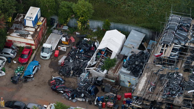 Dagenham United Kingdom 10 July 2026 Aerial view of a car salvage yard filled with piles of automotive parts scrap metal and stacked bumpers on scaffolding surrounded by industrial fencing.