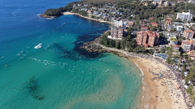 Aerial view of Manly Beach with swimmers in the turquoise water and people on the sandy shore surrounded by coastal buildings and lush greenery in Manly New South Wales Australia.