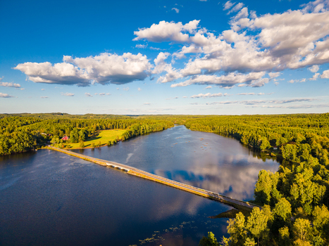 Aerial view of a bridge crossing a calm lake surrounded by lush green forests under a bright blue sky in Gallared Halland County Sweden. Aerial view of a bridge crossing a calm lake surrounded by lush green forests under a bright blue sky in Gallared Halland County Sweden.