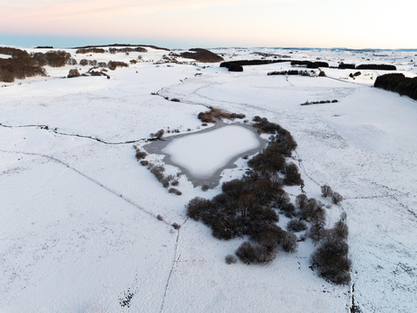 Aerial view of a serene snow blanketed landscape where a frozen lake mirrors the soft hues of the dawn sky Nasbinals Occitanie France.
