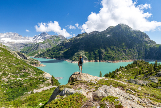 View of a hiker standing on a rock overlooking the turquoise waters of Lac d Emosson with the VerticAlp Emosson mountains in the background in Le Chatelard Valais Switzerland. View of a hiker standing on a rock overlooking the turquoise waters of Lac d Emosson with the VerticAlp Emosson mountains in the background in Le Chatelard Valais Switzerland.