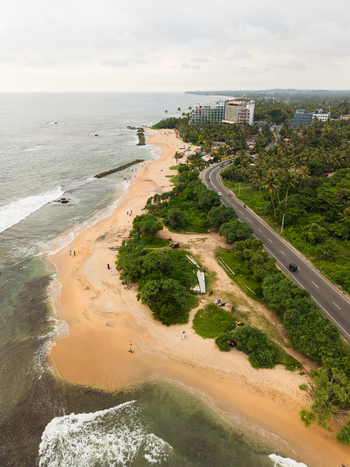 Aerial view of Dadalla beach featuring the sandy coastline a winding coastal road lush palm trees and modern buildings in Galle Southern Province Sri Lanka. Aerial view of Dadalla beach featuring the sandy coastline a winding coastal road lush palm trees and modern buildings in Galle Southern Province Sri Lanka.
