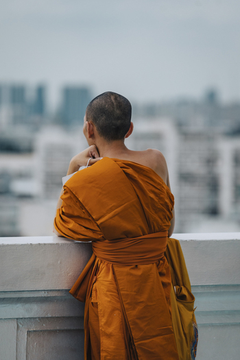 View of a serene monk in orange robe meditating on a balcony overlooking the modern cityscape Bangkok Thailand.