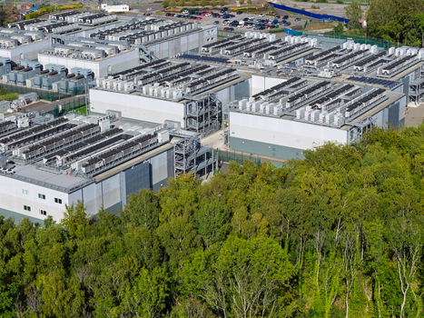 Aerial view of a large data center facility with multiple buildings and rooftop cooling units surrounded by dense forest in the United Kingdom.