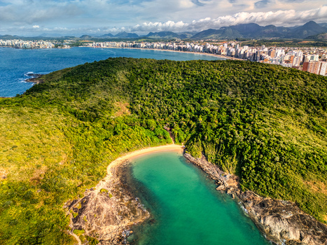 Aerial view of Praia do Ermitao featuring a secluded sandy beach nestled within lush green hills and rocky cliffs with the coastal city skyline in the distance Guarapari Espirito Santo Brazil. Aerial view of Praia do Ermitao featuring a secluded sandy beach nestled within lush green hills and rocky cliffs with the coastal city skyline in the distance Guarapari Espirito Santo Brazil.