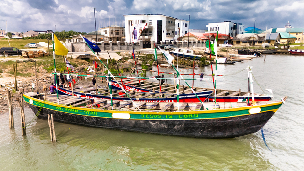 Aerial view of colorful wooden boats docked at a riverbank with flags nearby buildings and a cloudy sky in Minima Rivers Nigeria.
