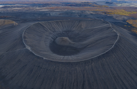 Aerial view of the stark grey black volcanic crater its textured slopes contrasting with the distant autumn tinged vegetation shapes a surreal landscape Myvatn &THORN;ingeyjarsveit Iceland.