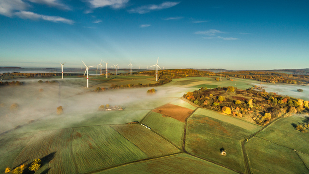 Aerial view of wind turbines standing in a rural landscape with agricultural fields patches of morning mist and autumn foliage under a clear blue sky in Germany. Aerial view of wind turbines standing in a rural landscape with agricultural fields patches of morning mist and autumn foliage under a clear blue sky in Germany.