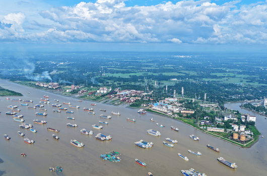 Aerial view of Karnaphuli River filled with cargo ships and barges industrial facilities and lush green banks under a cloudy blue sky Chattogram Chittagong Division Bangladesh.