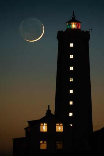 View of Grotta Lighthouse silhouetted against a twilight sky with a crescent moon and glowing windows in Seltjarnarnes Seltjarnarnesbaer Iceland. View of Grotta Lighthouse silhouetted against a twilight sky with a crescent moon and glowing windows in Seltjarnarnes Seltjarnarnesbaer Iceland.