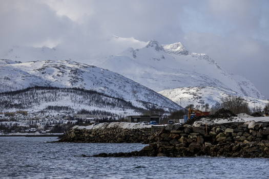 View of snow covered mountains rising above a rocky coastline and calm water with construction equipment on the shore in Kvaloya Troms Norway. View of snow covered mountains rising above a rocky coastline and calm water with construction equipment on the shore in Kvaloya Troms Norway.
