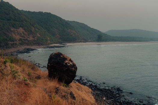View of a serene coastal landscape with a massive dark boulder perched on a grassy cliff edge overlooking the tranquil waters Chiplun Maharashtra India. View of a serene coastal landscape with a massive dark boulder perched on a grassy cliff edge overlooking the tranquil waters Chiplun Maharashtra India.