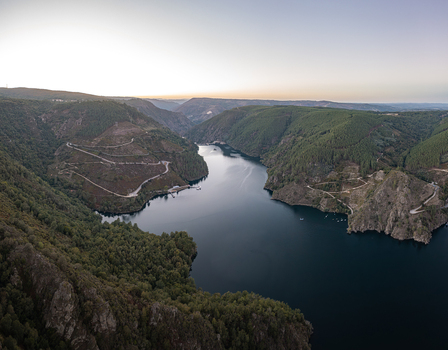 Aerial view of the Ribeira Sacra canyon with the Sil River winding through dense forests and steep hills under a soft sunset sky in Lugo Orense Spain. Aerial view of the Ribeira Sacra canyon with the Sil River winding through dense forests and steep hills under a soft sunset sky in Lugo Orense Spain.
