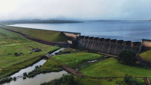 Aerial view of Midmar Dam featuring the concrete spillway and reservoir surrounded by green hills under a misty sky in Howick KwaZulu Natal South Africa. Aerial view of Midmar Dam featuring the concrete spillway and reservoir surrounded by green hills under a misty sky in Howick KwaZulu Natal South Africa.