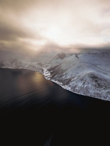 Aerial view of where the dark waters meet the snow covered peaks under a luminous sky Fjordgård Troms Norway. Aerial view of where the dark waters meet the snow covered peaks under a luminous sky Fjordgård Troms Norway.
