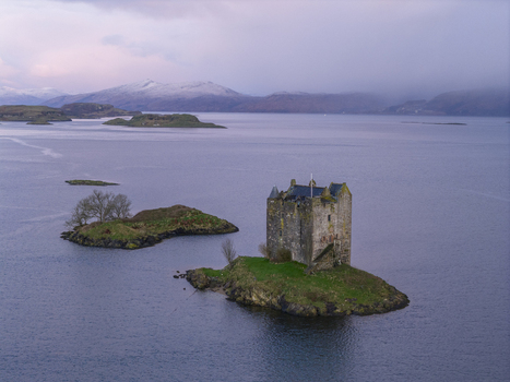 Aerial view of Castle Stalker on a small green islet surrounded by calm water and snow capped mountains in the background Appin Scotland United Kingdom. Aerial view of Castle Stalker on a small green islet surrounded by calm water and snow capped mountains in the background Appin Scotland United Kingdom.
