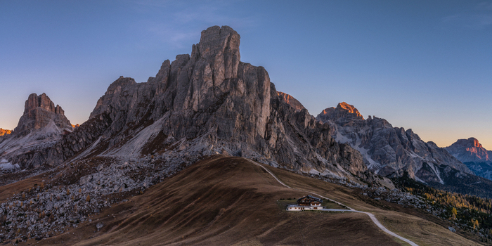 View of the dramatic limestone peaks of the Ra Gusela in the Dolomites with a mountain hut nestled on the grassy slopes under a clear sky at sunset in Italy.