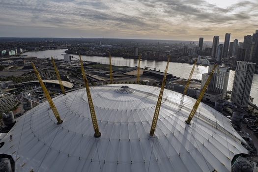 Aerial view of The O2 Arena with its iconic yellow masts surrounded by the River Thames and the London skyline under a cloudy sky in London England United Kingdom. Aerial view of The O2 Arena with its iconic yellow masts surrounded by the River Thames and the London skyline under a cloudy sky in London England United Kingdom.