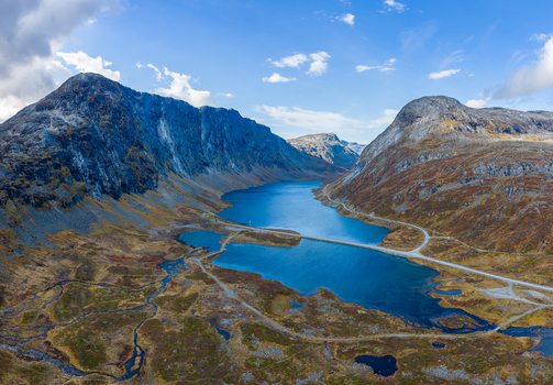 Aerial view of Skjakvegen winding through the rugged mountain landscape alongside a deep blue lake with autumn foliage in Skjak Innlandet Norway. Aerial view of Skjakvegen winding through the rugged mountain landscape alongside a deep blue lake with autumn foliage in Skjak Innlandet Norway.