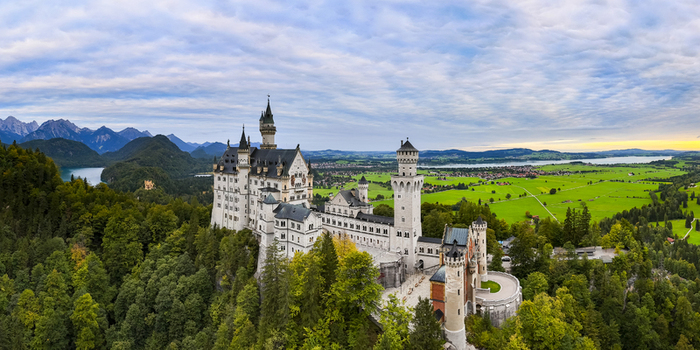 Aerial view of the majestic Neuschwanstein Castle piercing the sky above a sea of emerald trees with distant mountains framing the horizon Neuschwanstein Castle Bayern Germany. Aerial view of the majestic Neuschwanstein Castle piercing the sky above a sea of emerald trees with distant mountains framing the horizon Neuschwanstein Castle Bayern Germany.