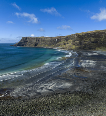 Aerial view of Talisker Bay Beach with its dark sand turquoise waves and a waterfall cascading down the rugged cliffs under a blue sky in Isle of Skye Scotland United Kingdom. Aerial view of Talisker Bay Beach with its dark sand turquoise waves and a waterfall cascading down the rugged cliffs under a blue sky in Isle of Skye Scotland United Kingdom.