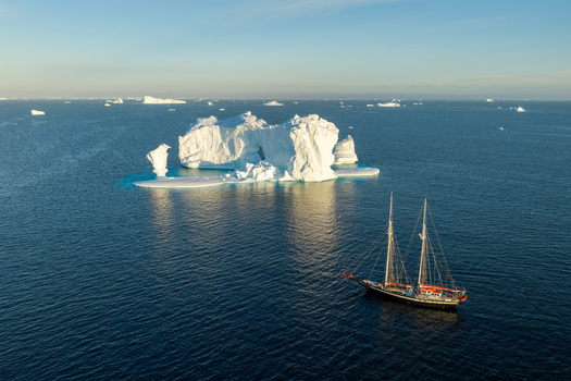 Aerial view of a majestic arch iceberg floating in the deep blue waters of the Arctic Ocean with a classic sailing ship passing by in Greenland. Aerial view of a majestic arch iceberg floating in the deep blue waters of the Arctic Ocean with a classic sailing ship passing by in Greenland.