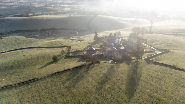 Aerial view of farmhouses nestled in rolling green hills with long shadows stretching across the landscape bathed in the soft golden light of early morning England United Kingdom. Aerial view of farmhouses nestled in rolling green hills with long shadows stretching across the landscape bathed in the soft golden light of early morning England United Kingdom.