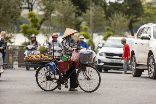 Hanoi Vietnam 11 November 2025 View of a street vendor wearing a conical hat riding a bicycle loaded with baskets of potatoes on a busy city street with motorbikes.