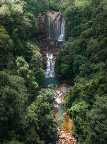 Aerial view of a waterfall cascading through a dense tropical rainforest with lush green trees and rocky riverbed Costa Rica.