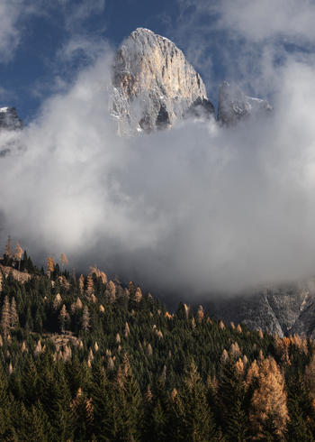 View of the Dolomites mountain peak partially shrouded in thick white clouds above an autumn forest with yellow and green trees Trentino Alto Adige Italy. View of the Dolomites mountain peak partially shrouded in thick white clouds above an autumn forest with yellow and green trees Trentino Alto Adige Italy.