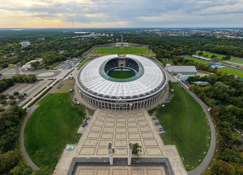 Berlin Germany 26 September 2026 Aerial view of Olympiastadion Berlin with its iconic roof structure surrounding green parklands and paved plaza under a soft evening sky.