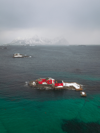Aerial view of a vibrant red fishing cabin stands out against the dark frigid sea and snow dusted rocks with snowy mountains in the distance Svolv&aelig;r Nordland Norway.