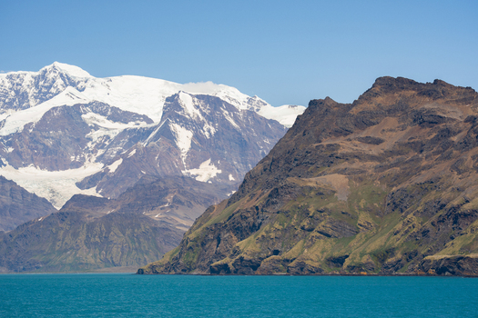 View of the rugged coastal mountains and snow capped peaks rising above the turquoise waters of the bay in Grytviken South Georgia and the South Sandwich Islands. View of the rugged coastal mountains and snow capped peaks rising above the turquoise waters of the bay in Grytviken South Georgia and the South Sandwich Islands.