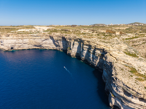 Aerial view of the steep limestone cliffs at Dwejra Bay overlooking the deep blue Mediterranean sea with a small boat and rocky terrain in Gozo Region Malta. Aerial view of the steep limestone cliffs at Dwejra Bay overlooking the deep blue Mediterranean sea with a small boat and rocky terrain in Gozo Region Malta.