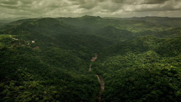 Aerial view of lush green mountains and dense tropical forest with a winding river flowing through the valley under a cloudy sky in San Sebastian San Sebastian Puerto Rico.
