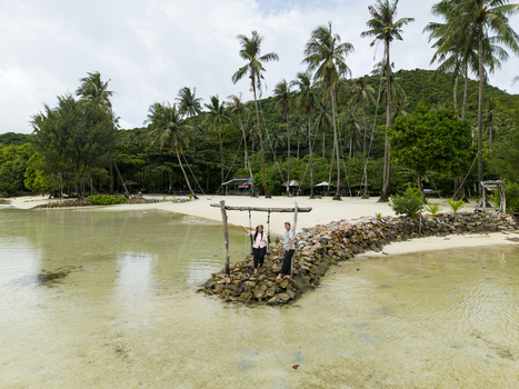 Aerial view of a couple standing on a rock formation under a wooden swing framed by palm trees and the serene waters of Bobby Beach Karimunjawa Central Java Indonesia.