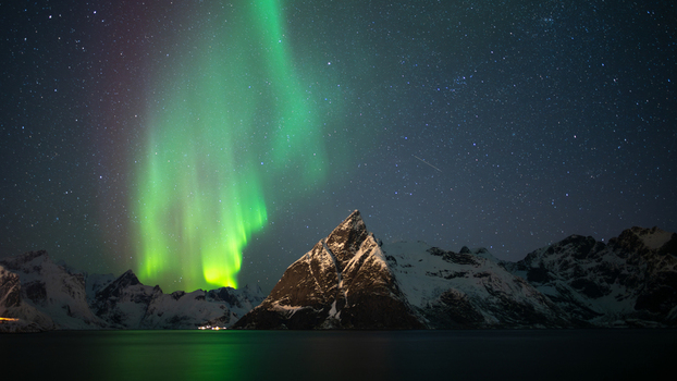 View of the vibrant green aurora borealis illuminating the night sky above the snow capped mountain peaks and dark waters in Leknes Lofoten Norway.