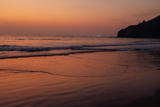 View of the tranquil beach reflecting the fiery sunset as gentle waves kiss the shore near the dark silhouette of a distant headland Chiplun Maharashtra India. View of the tranquil beach reflecting the fiery sunset as gentle waves kiss the shore near the dark silhouette of a distant headland Chiplun Maharashtra India.