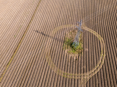 Aerial view of a high voltage electricity pylon standing in the center of a large plowed agricultural field with distinct circular patterns in the furrows in Poland.