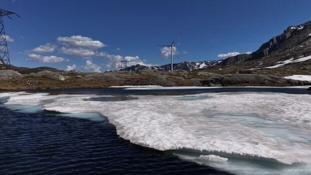 Aerial view of San Gottardo wind Park with a wind turbine standing near a partially frozen mountain lake under a clear blue sky in Airolo Ticino Switzerland.